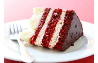 A delicious-looking slice of layered red velvet cake topped with white frosting sitting on a plate with a fork, ready to eat.