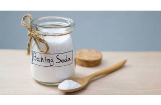 A small glass jar of baking soda sitting on a table. Next to it is a cork lid and a wooden spoon containing baking soda.